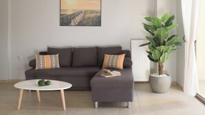 Een woonkamer met een grijze bank, twee gekleurde kussens, een witte bijzettafel met avocado's, een grote potplant en een ingelijste foto aan de muur.