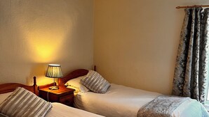 A hotel room with two single beds, a nightstand with a lamp, a wooden headboard, and a window with floral curtains. - 🌿 Peaceful & Cosy Cottage Stay in the Forest of Dean 🌳🐑 (Lydney)