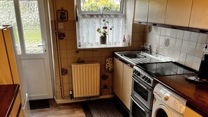 A compact kitchen with wooden flooring, a built-in oven, a washing machine, and a window with a view of greenery. - 🌿 Peaceful & Cosy Cottage Stay in the Forest of Dean 🌳🐑 (Lydney)