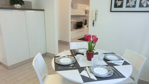 En spiseplads med et rundt bord dækket til fire personer, hvide stole og en vase med lyserøde blomster.
