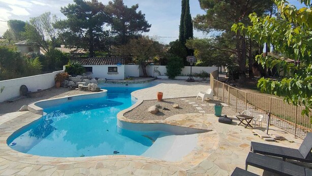 A swimming pool with a stone deck, surrounded by a fence, trees, and a white building in the background. - Cottage with private garden in wooded property with swimming pool (Pertuis)