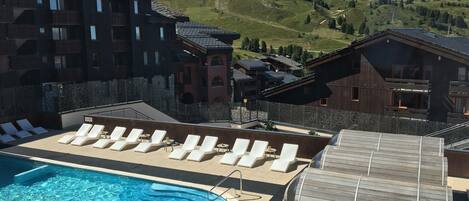 A pool area with lounge chairs and a wooden barrel, surrounded by buildings with balconies and a mountain backdrop.