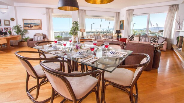 A spacious living area with a glass dining table, cushioned chairs, and a sofa. There are large windows offering a view of the city.
