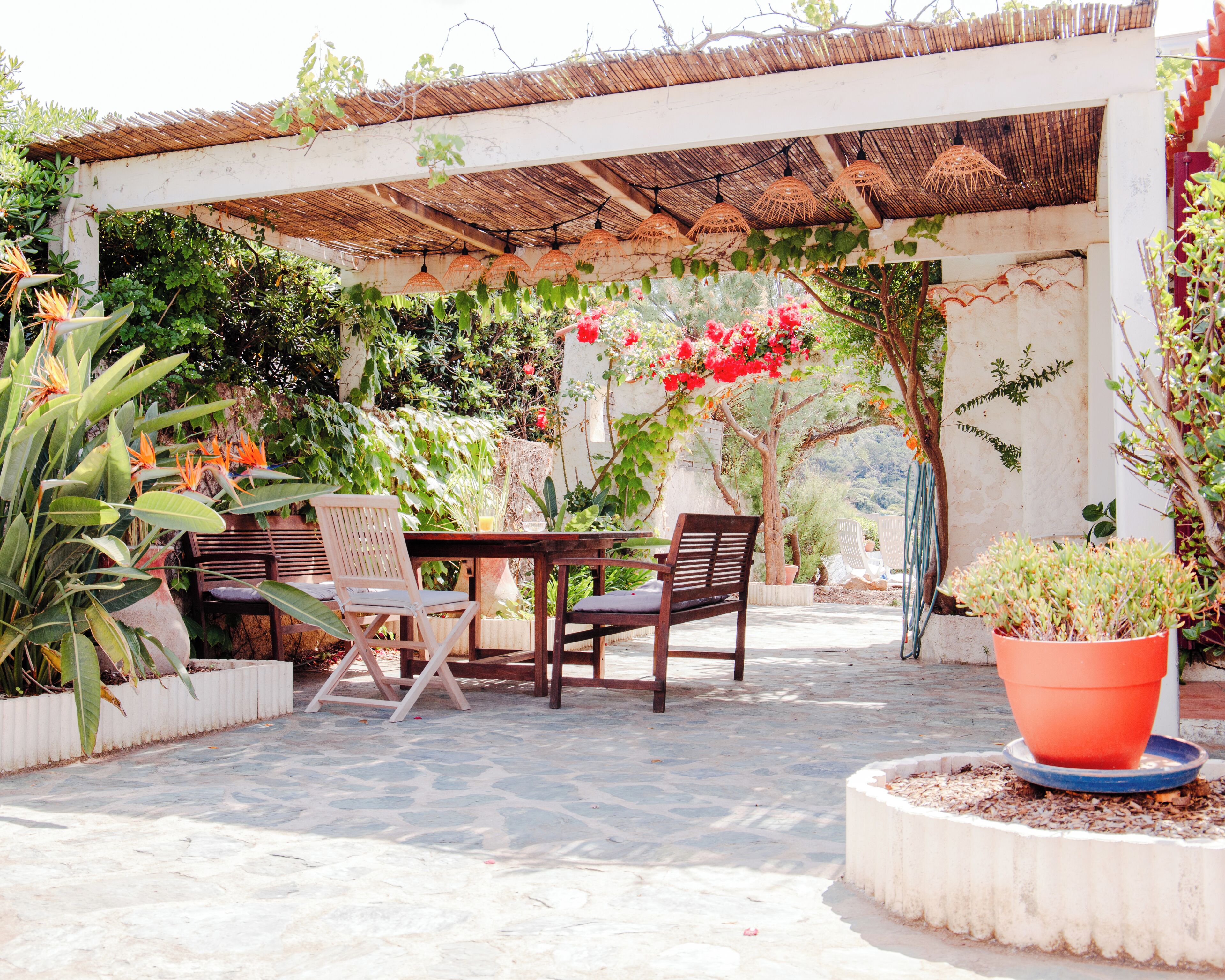 A covered outdoor seating area with a table and chairs, surrounded by potted plants and a pergola with hanging lights.