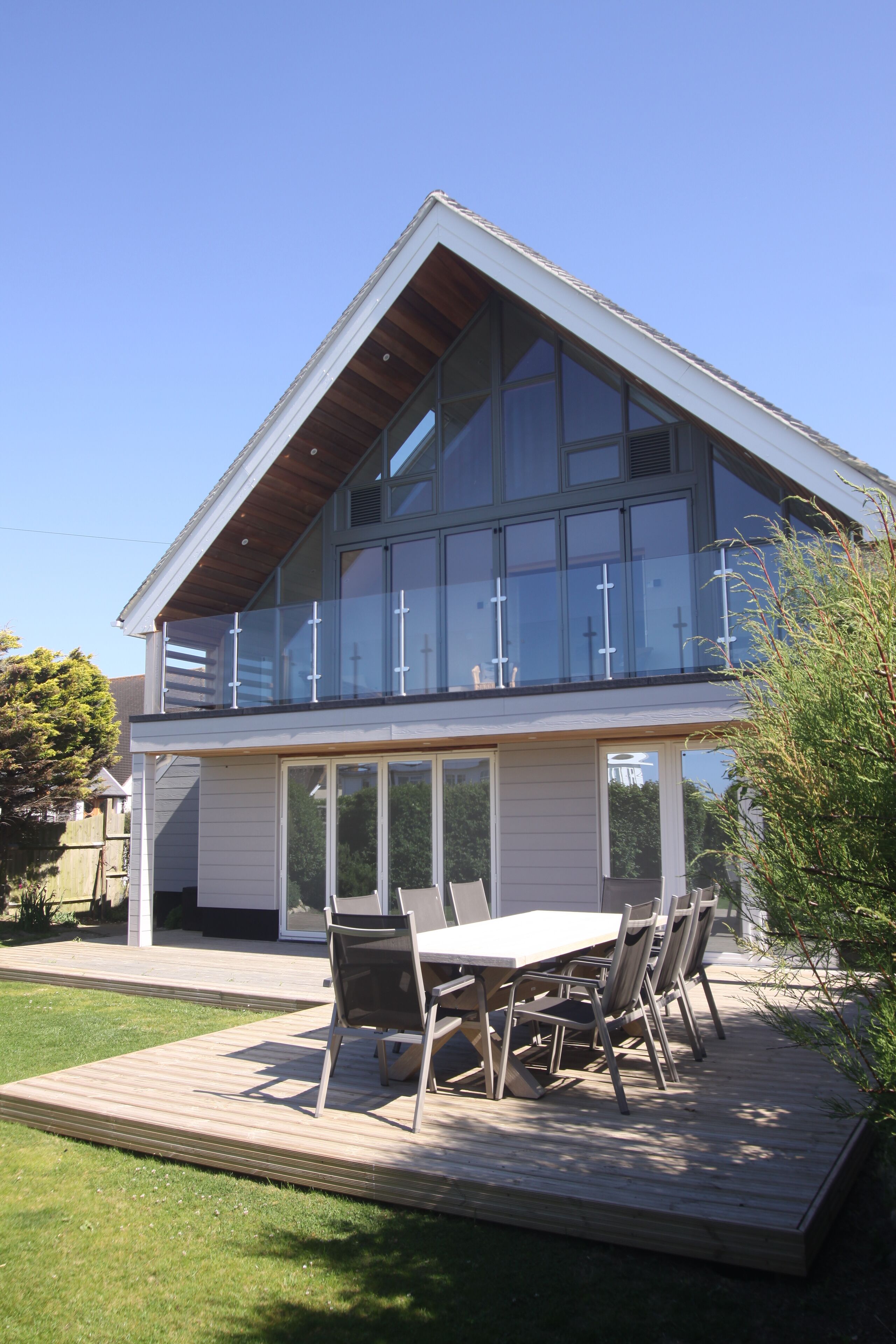 Une maison moderne à deux étages, dotée d’un balcon, de grandes fenêtres et d’une terrasse aménagée avec du mobilier d’extérieur.