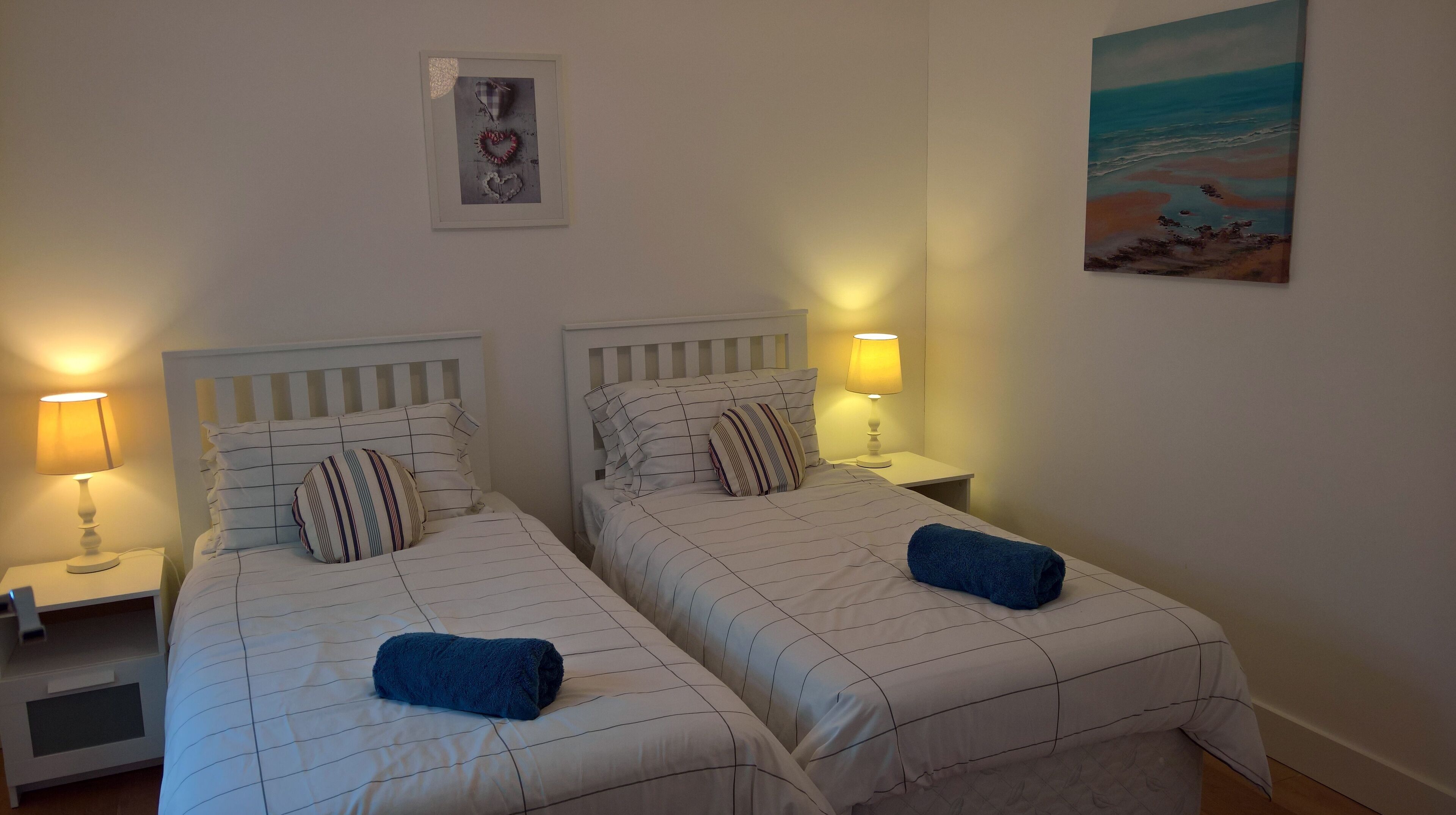 Habitación de hotel con dos camas, cada una con una almohada a rayas y una toalla azul. Hay lámparas de noche y un cuadro en la pared.