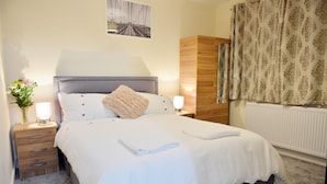 A neatly made bed with white linens, a bedside lamp, and a nightstand with drawers. - 3 bedroom London House (Dagenham)