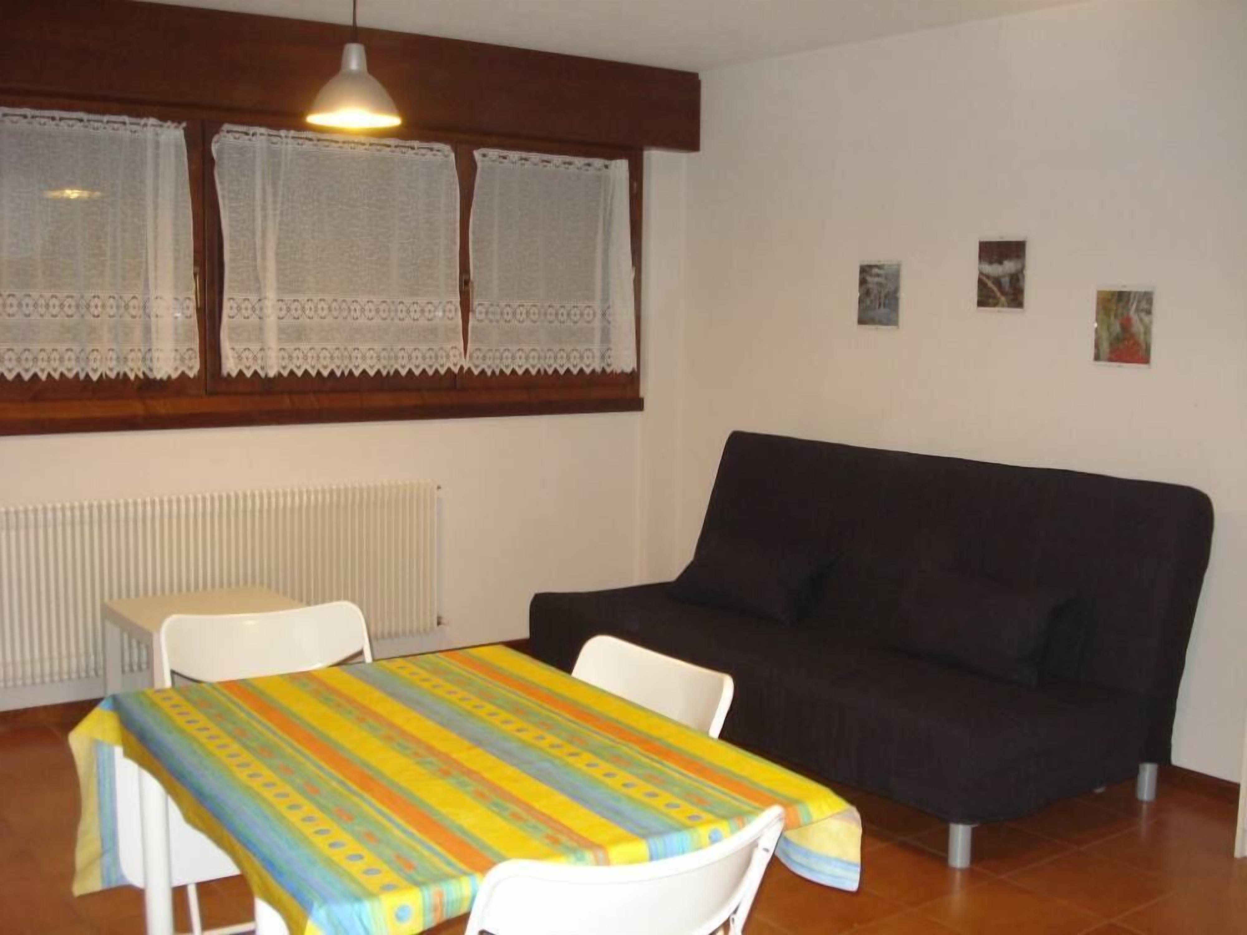 A room with a dark sofa, a white table with a colorful tablecloth, and a window with lace curtains.