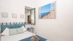 Ein ordentlich eingerichtetes Schlafzimmer mit einem Bett, Kissen, einer blauen Bettdecke, einem gefalteten Handtuch, einem kleinen Tisch mit einer Lampe und einem Bilderrahmen mit einem Strandbild.