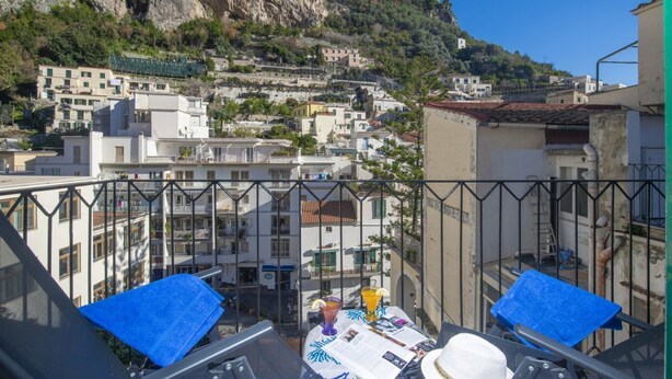 Ein Balkon mit Tisch, einem weißen Hut und Blick auf eine Hügelstadt mit Gebäuden.