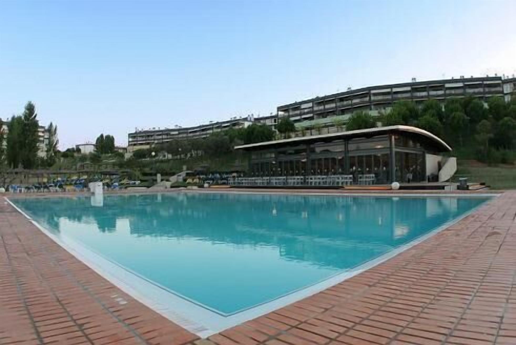 A rectangular swimming pool with a brick border, surrounded by a building with a curved roof and a balcony overlooking the pool.