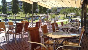 An outdoor patio area with wooden chairs and tables, umbrellas, and a view of a pool and greenery in the background.
