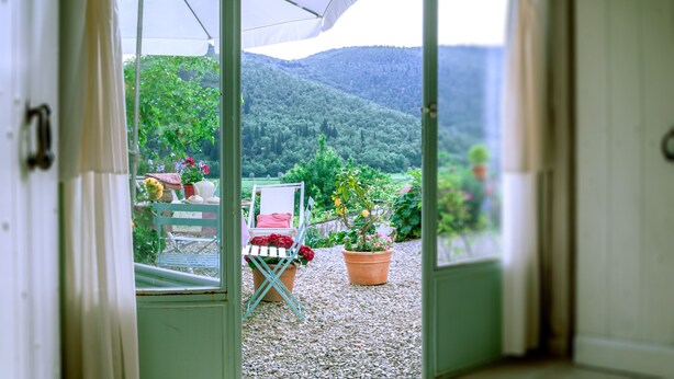 Udsigt fra åbent dør ud til en patio med spisebord, omgivet af krukkeplanter og en stol.