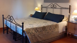 Uma cama com colcha estampada, duas almofadas, mesa de cabeceira com abajur e um quadro na parede.