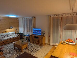 A living room with a sofa, a coffee table, a TV, a lamp, and a dining table with fruit.