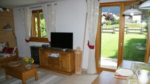 Ein Wohnzimmer mit Fernseher, Holzmöbeln, einem Esstisch mit einer Karaffe und Blick auf eine grüne Landschaft durch eine Glastür.