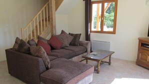 Un salon avec un canapé marron, un pouf et une table basse en bois. On aperçoit un escalier en bois à l’arrière-plan.