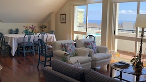A spacious living area with a dining table, two sofas, and a view of the sea through large windows.