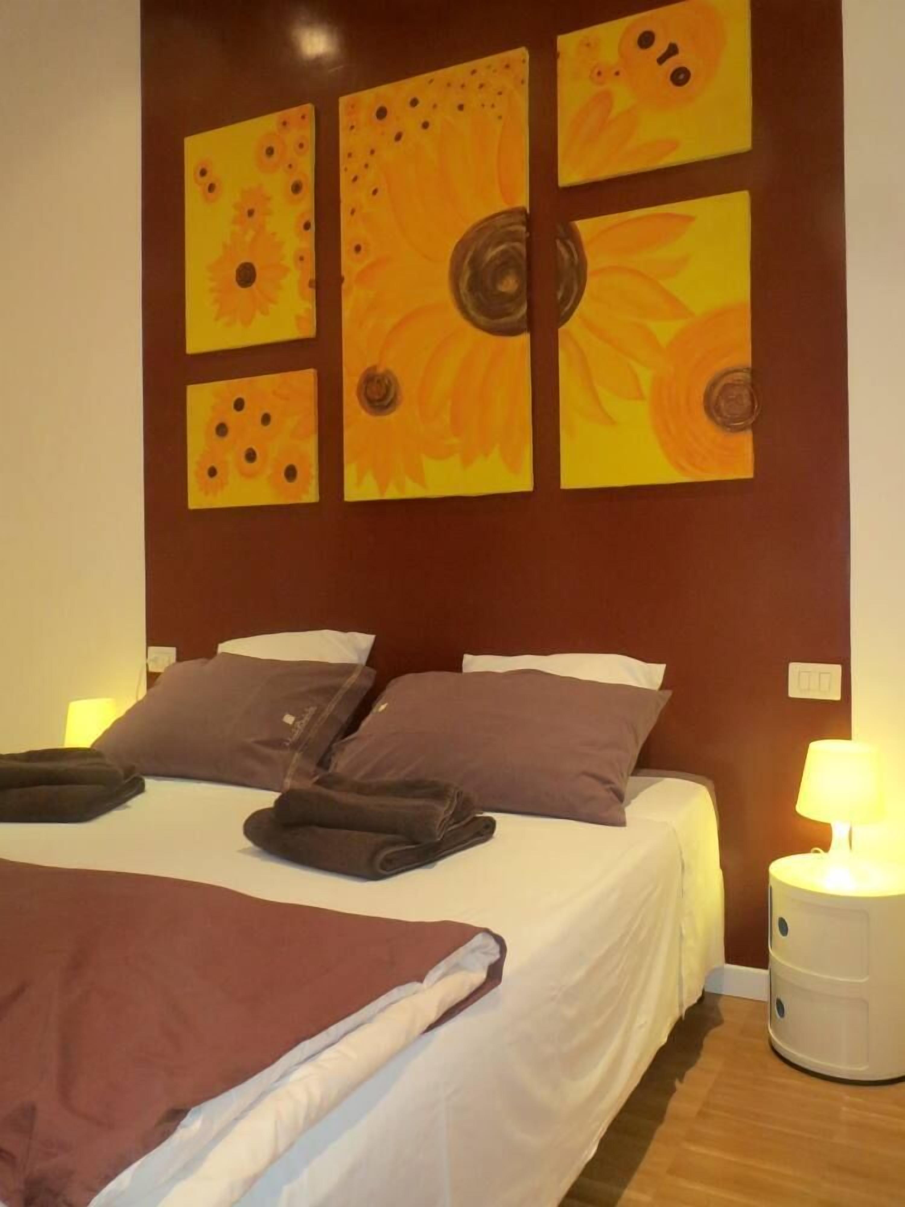 A hotel room with a bed, pillows, a blanket, and artwork on the wall.