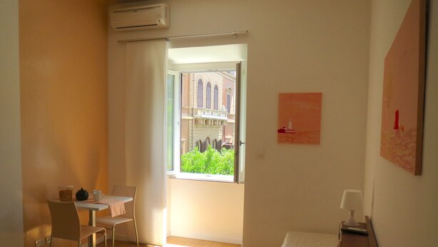 A hotel room with a bed, a desk, a chair, a table, and a window with a view of buildings and greenery. - Rome: NEW from 3 up to 11 people Central, Modern and bright (Roma)
