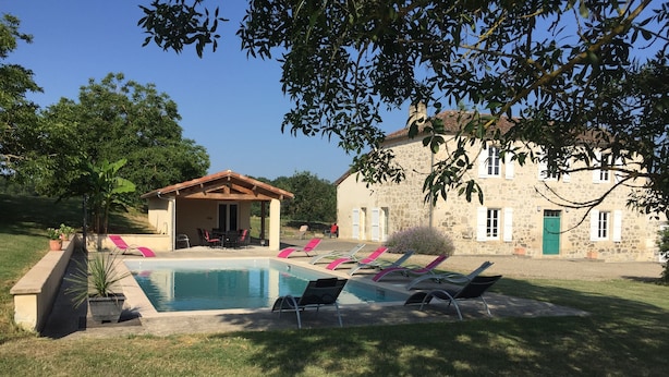 Zona de piscina con sillones reclinables, un espacio de estar techado y un edificio de piedra al fondo.