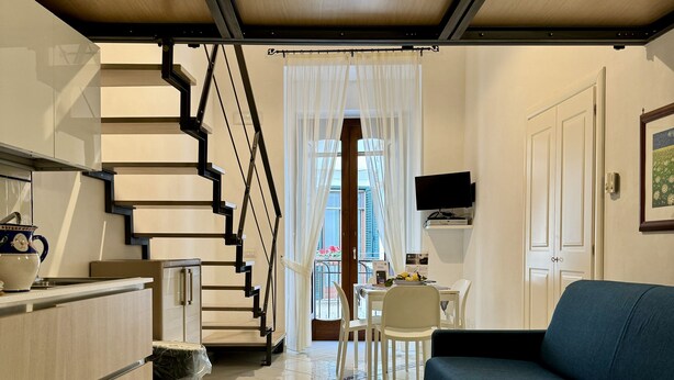 A modern living room with a blue sofa, a kitchen area with a stove, and a staircase leading to a balcony.