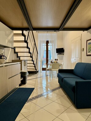 A modern living room with a blue sofa, a kitchen area with a stove, and a staircase leading to a balcony.