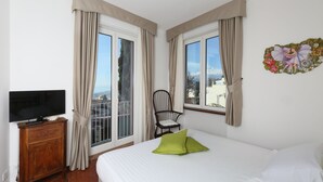 A hotel room with a bed, a television on a wooden cabinet, a chair, and large windows with curtains.