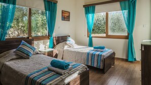 A room with two beds, each with a striped blanket and a pillow, a bedside table with a lamp, and a window with teal curtains. - Villa Astro -  a beach villa with heated pool that sleeps 8 guests  in 4 bedrooms (Paphos)