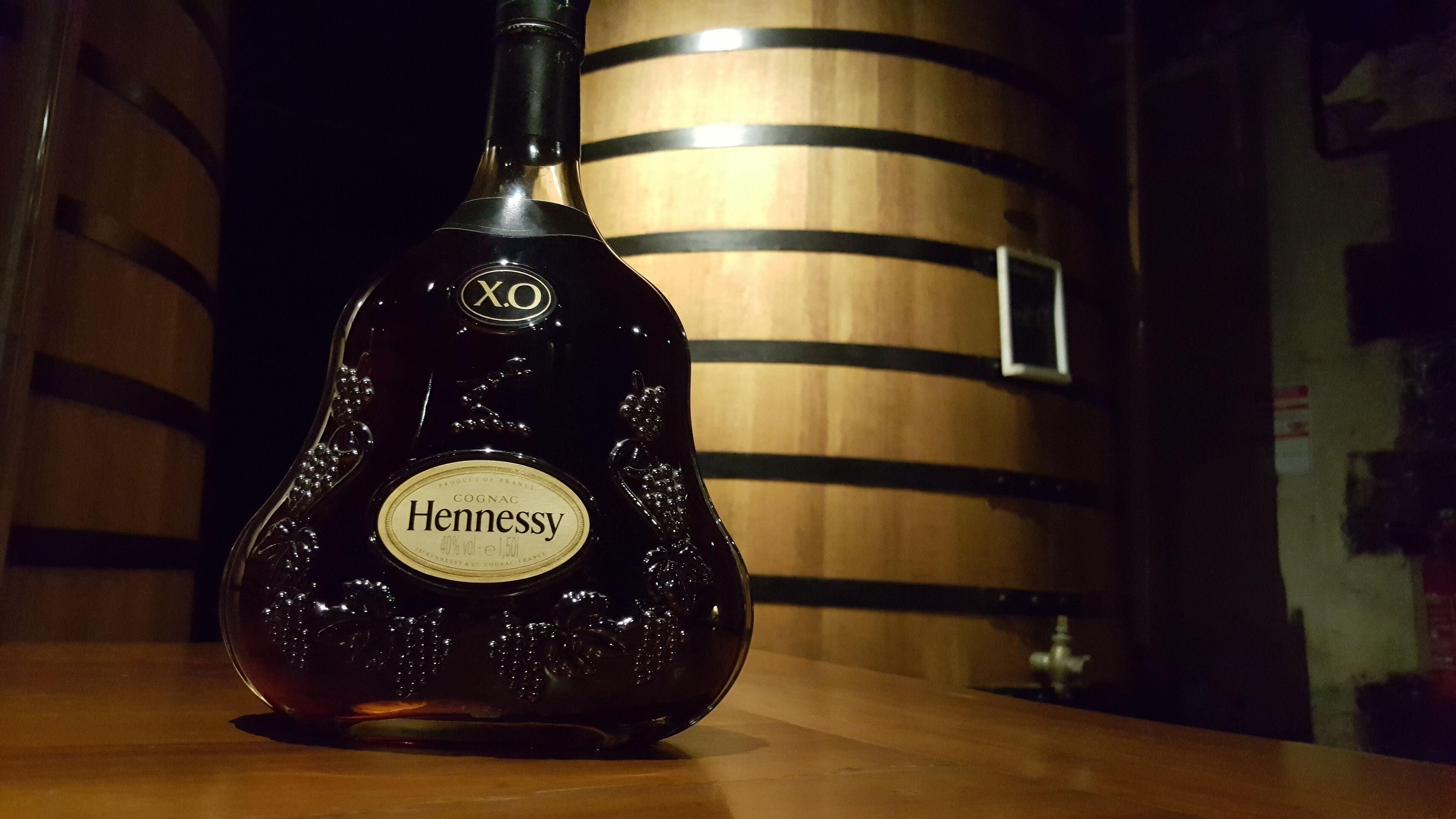 A bottle of Cognac Hennessy on a wooden surface with a barrel in the background.