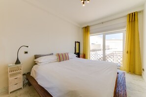 A bedroom with a large bed, bedside tables, a nightstand, a window with yellow curtains, and a view of the outside.