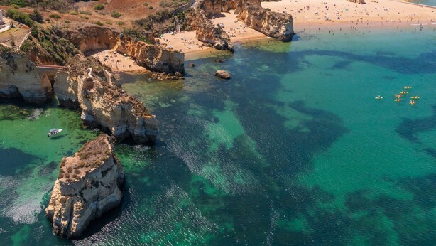 Una vista aerea di una zona costiera con spiaggia, scogliere rocciose e acque cristalline color turchese.