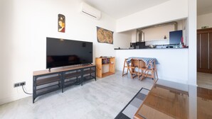 Un soggiorno moderno con una TV a schermo piatto, un mobile basso in legno, un piccolo tavolo da pranzo con sedie e un angolo cottura con lavello e piano di lavoro.