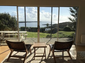 A room with a large sliding glass door leading to a patio with a table and two chairs, overlooking a body of water and greenery. - Private waterfront house with its own 3 acres sloping to the sea (Tralispean)