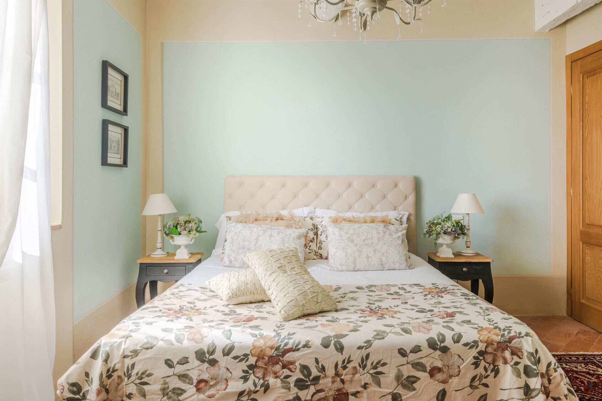 A bedroom with a floral bedspread, a tufted headboard, two bedside tables with lamps, and framed pictures on the wall.