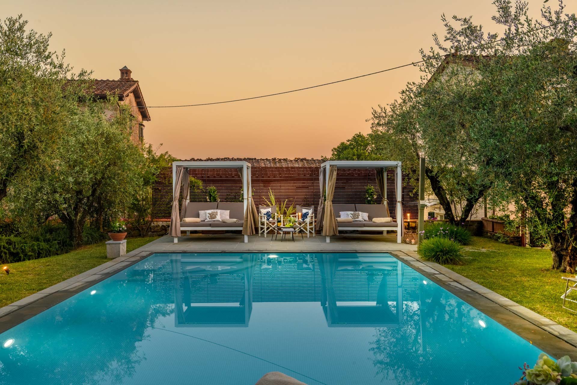 A swimming pool with two outdoor seating areas, surrounded by greenery and a building in the background.