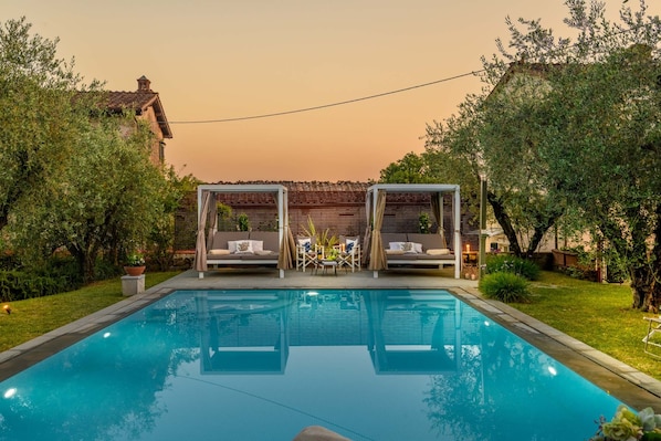 A swimming pool with two outdoor seating areas, surrounded by greenery and a building in the background.