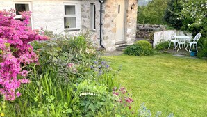 A stone house with a sloped roof, a small garden with various plants and flowers, and a white outdoor table and chairs. - Stunning views of Snowdonia Mountains (Near Caernarfon)