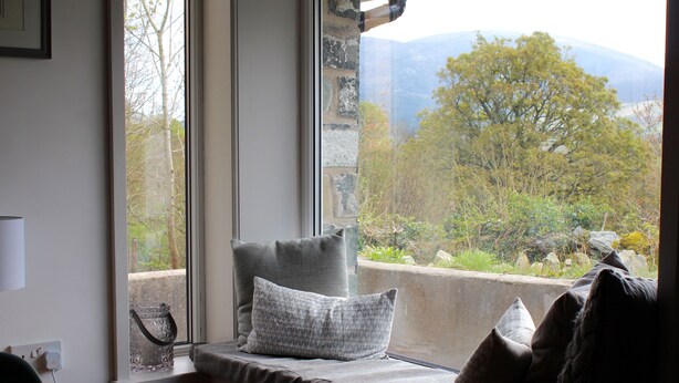 A room with a large window overlooking a scenic landscape, a cushioned bench with pillows, and a wooden table. - Stunning views of Snowdonia Mountains (Near Caernarfon)