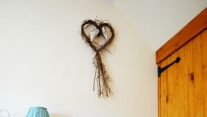 Une pièce d’angle avec un fauteuil en osier, une petite table en bois surmontée d’une lampe et un élément décoratif en forme de cœur accroché au mur.