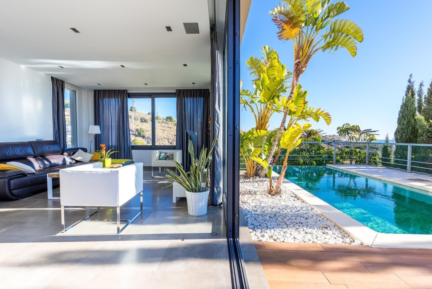 A modern living room with a sofa, coffee table, and potted plants. There is a large window with curtains and a view of a swimming pool outside.