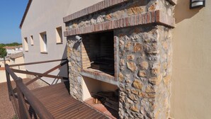Un barbecue en pierre installé à l’extérieur, adossé à un bâtiment, avec une terrasse en bois et une rambarde en métal.