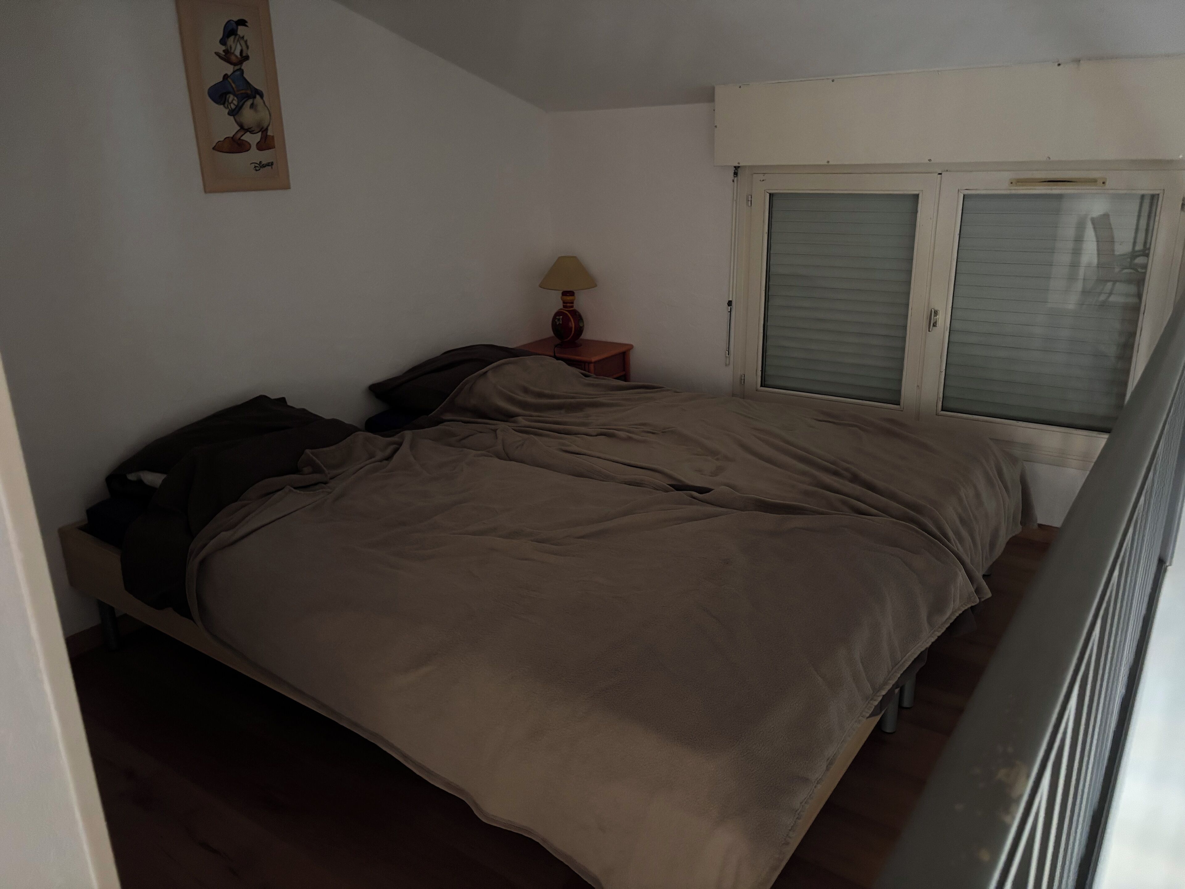 A bedroom with a bed, a nightstand with a lamp, a window with blinds, and a wall decoration.