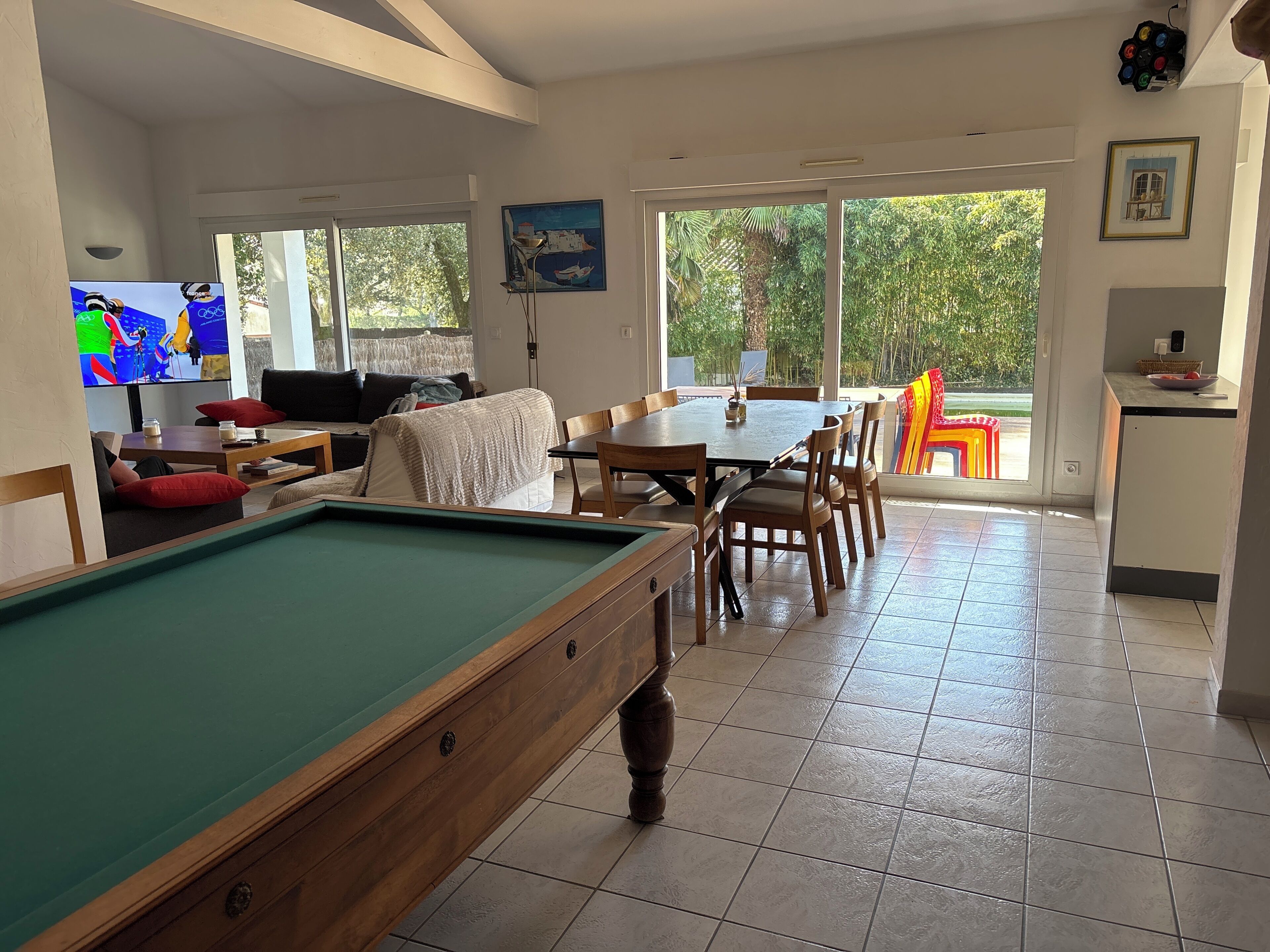Une table de billard dans une pièce bien éclairée, avec un coin repas et de grandes fenêtres.