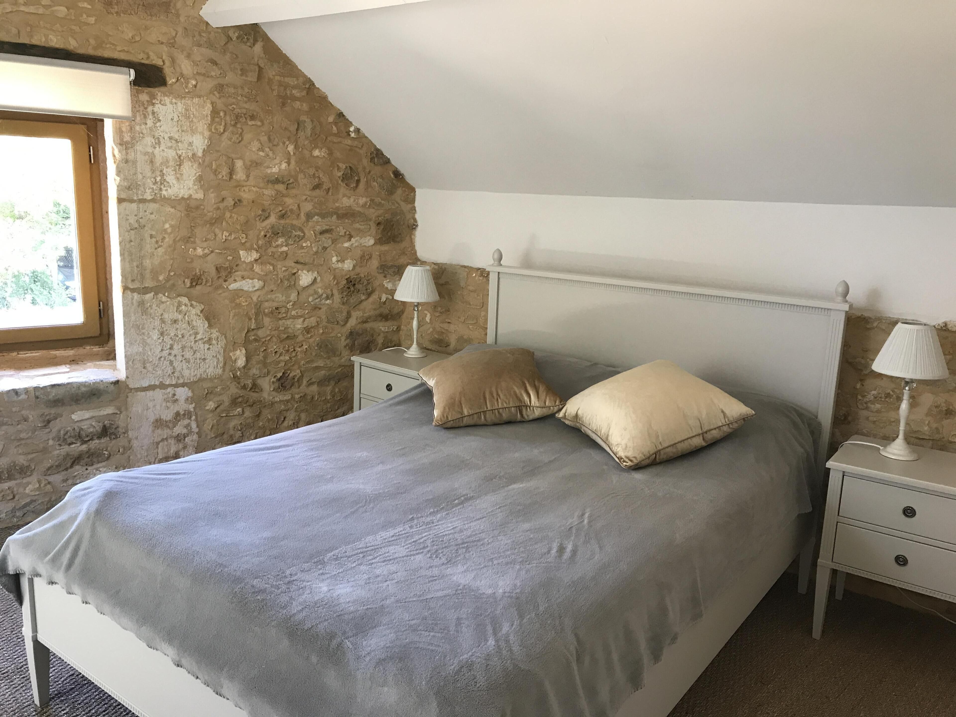 Ein Schlafzimmer mit Steinwand, schrägen Decken, einem Bett mit grauer Decke, zwei Nachttischlampen und einem Fenster mit Blick auf Grünflächen.