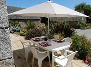 Une table blanche, quatre chaises et un grand parasol, installés sur une terrasse aux murs de pierre et agrémentée de plantes.