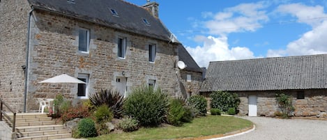 A stone building with a grey roof, a small garden with plants and a white chair, and a gravel driveway.