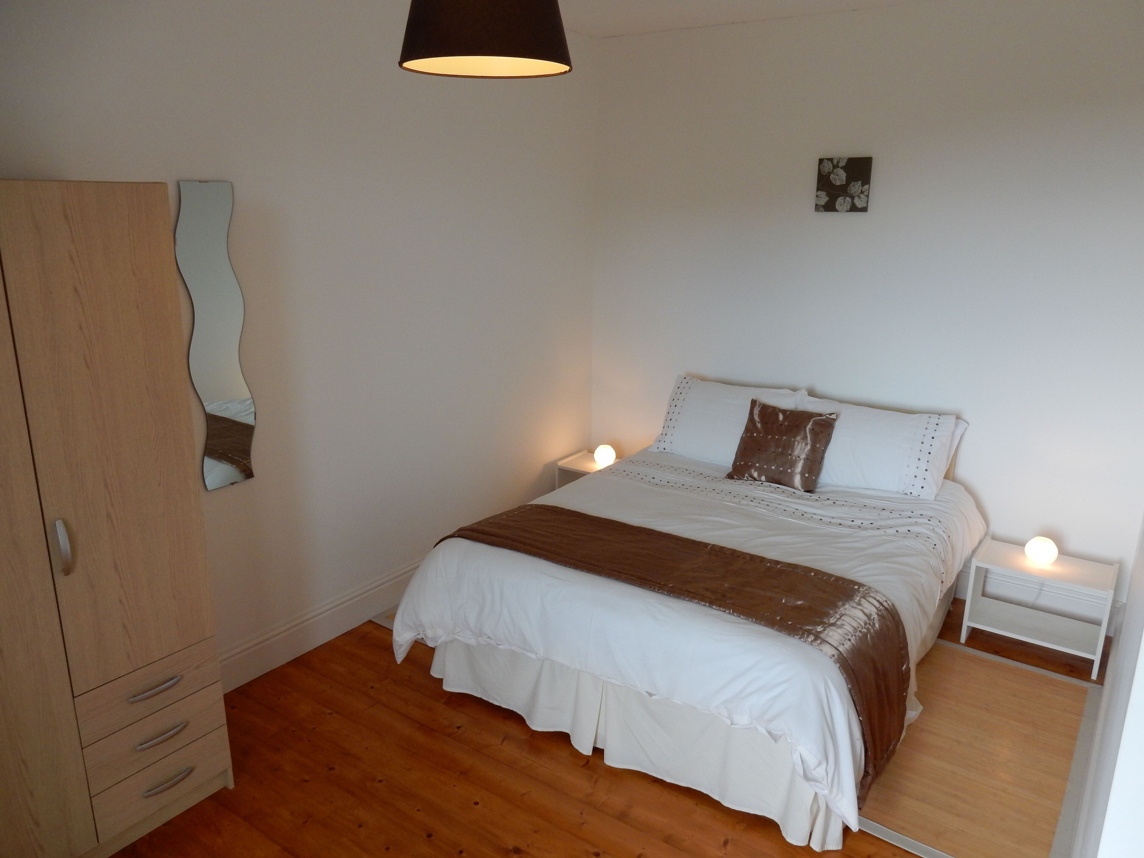 A bedroom with a wooden wardrobe, a bed with white bedding and a brown throw, a small bedside table with a lamp, and a wall-mounted mirror.
