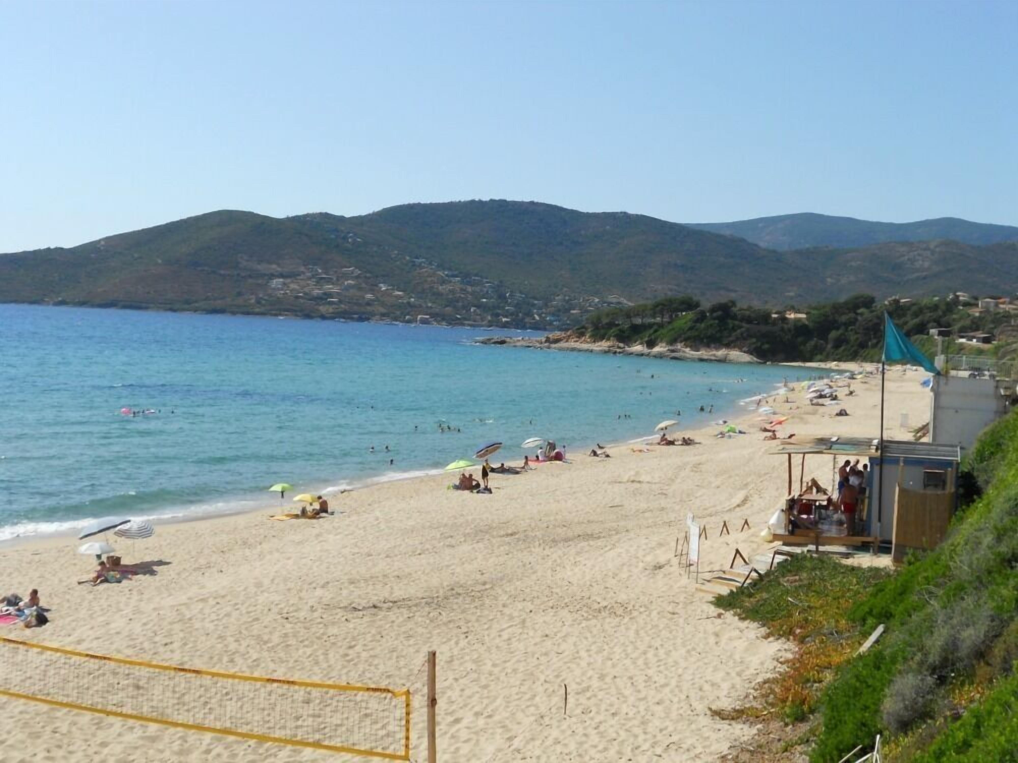 Ein Strand mit klarem, blauem Wasser, sandigem Ufer und Hügeln im Hintergrund.