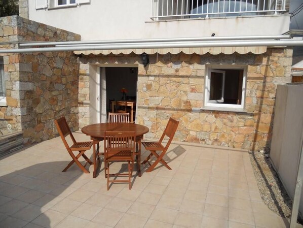 Una casa de piedra con un patio empedrado, una mesa de madera con cuatro sillas y una ventana pequeña.